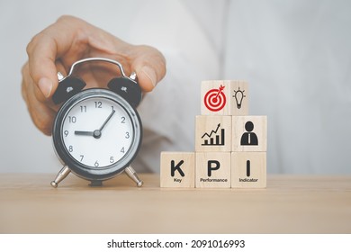 KPI, Key Performance Indicator, on wooden cube and business icon above with senior's  hand holding black analog alarm clock