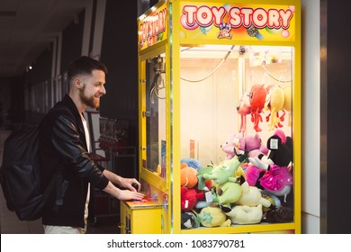 KRAKOW / POLAND - 05.02.2018: Young hipster man with a beard in black jacket and with backpack playing in a vending machine pullout soft toys. Strong an with beard playing in toys machine and smiling.