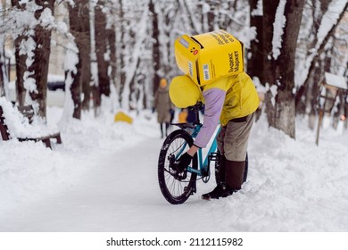 Krasnodar, Russia - January 24 2022: Courier, A Food Delivery Man With A Thermal Bag On His Back Repairing A Bicycle In The Winter Outside. Yandex Delivery.