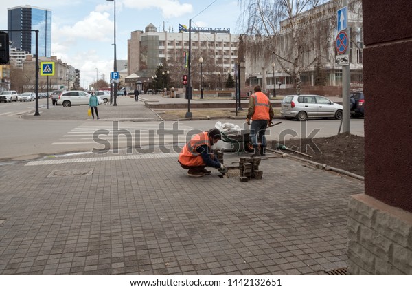 Krasnoyarsk,\
Krasnoyarsk Region / RF - April 25, 2019: Two workers in bright\
protective vests repair paving slabs in front of a pedestrian\
crossing on the background of a city\
street.