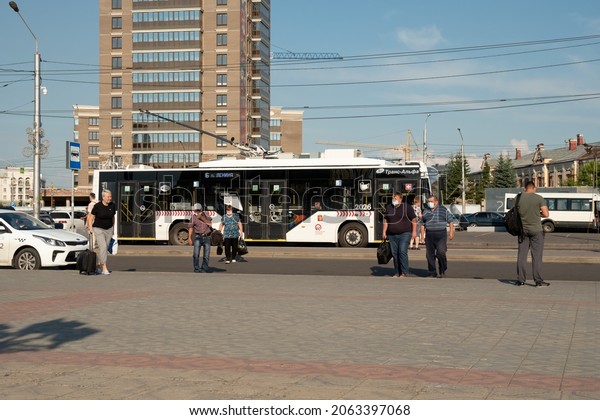 Krasnoyarsk, Krasnoyarsk Region, RF - July 11, 2021:\
People in medical masks with bags walk from the bus stop from the\
trolleybus past a taxi against the backdrop of city houses on a\
sunny summer day.