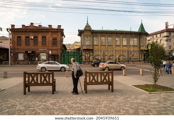 Krasnoyarsk, Krasnoyarsk Region, RF - July 19,\
2021: A gray-haired man with a stick and a bag over his shoulder\
stands near the benches on the pavement opposite the old houses on\
Karl Marx Avenue.