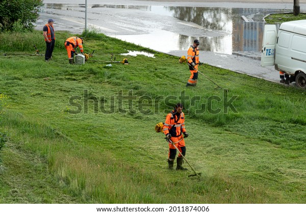 Krasnoyarsk, Krasnoyarsk Region, RF - June 29, ,\
2021: A group of workers in orange overalls mow the lawn on a\
cloudy summer day.