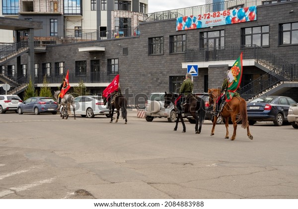 Krasnoyarsk,\
Krasnoyarsk Region, RF - May 9, 2021: Four Cossacks with flags in\
their hands ride horses past parked cars in the courtyard of a\
residential building in the spring\
city.