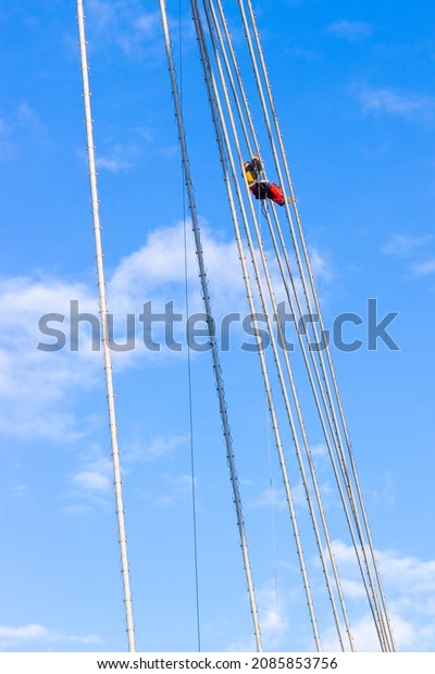 Krasnoyarsk, Russia -\
October 28, 2021: an industrial climber hangs on ropes and repairs\
lighting equipment installed on the cables of the cable-stayed\
bridge on a sunny\
day
