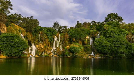 Kravice waterfall with multiple streams flowing over mossy cliffs into the calm green river, surrounded by lush vegetation, Bosnia and Herzegovina.