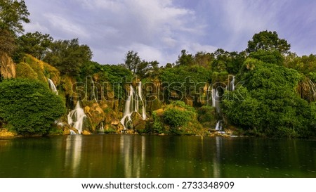 Kravice waterfall with multiple streams flowing over mossy cliffs into the calm green river, surrounded by lush vegetation, Bosnia and Herzegovina.