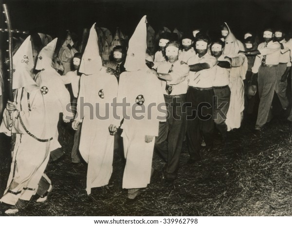 Ku Klux Klan initiation at Stone\
Mountain near Atlanta, Georgia. June 1949. KKK men in full white\
masks and gowns lead new members wearing small face\
masks