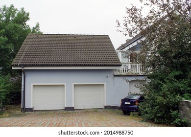 KUCHAROVICE, CZECH REPUBLIC - AUGUST 17, 2015. Private Blue Garage With Roller Shutters For Two Cars. The Garage Is Connected To The Main House By An Extension. A Dark Blue Car Is Parked Nearby