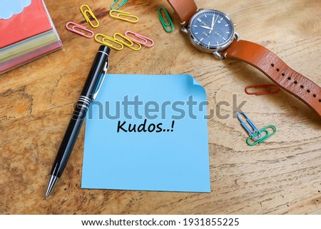 Kudos message written on the piece of paper, with a pen, paper clips, wristwatch and stacked of paper notes