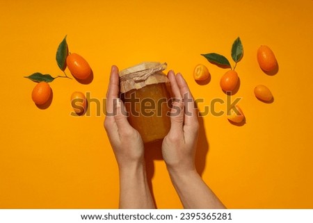 Kumquat and jar with jam in hands on orange background, top view