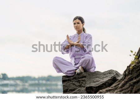 Kungfu master woman wearing kimono, practicing wushu sitting in lotus pose on stone near water, copy space. Healthy lifestyle, martial arts concept 