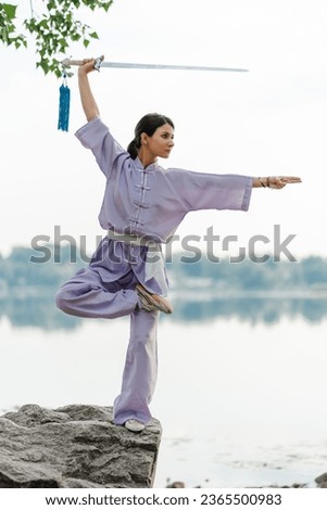 Kungfu master woman wearing kimono holding sword, practicing wushu standing on stone near water, copy space. Healthy lifestyle, martial arts concept 