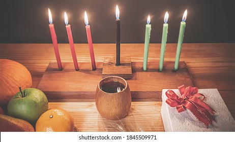 Das Urlaubskonzept von Kwanzaa mit sieben Kerzen in Rot, Schwarz und Grün, Geschenkbox, Kürbis, Mais und Obst auf Holzschreibtisch und schwarzem Hintergrund.