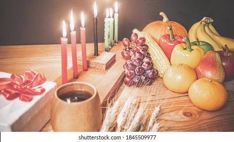 Das Urlaubskonzept von Kwanzaa mit sieben Kerzen in Rot, Schwarz und Grün, Geschenkbox, Kürbis, Mais und Obst auf Holzschreibtisch und schwarzem Hintergrund.