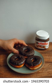 KYIV, UKRAINE - APRIL 1, 2021: Chocolate Donuts And A Can Of Nutella. Hand Holds A Chocolate Donut. Reach Out For A Donut With Nutella.