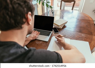 KYIV, UKRAINE - APRIL 13, 2020: Back View Of Young Man Writing On Paper While Using Laptop With Blank Screen