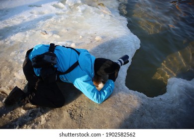 Kyiv, Ukraine - January, 19, 2021: Woman Photojournalist With Professional Photo Camera Knelt Down And Photographs Ice And Water In Winter