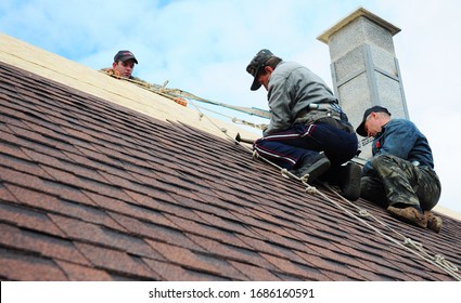KYIV, UKRAINE - MARCH, 29, 2020: Roofer contractors are installing dimensional asphalt shingles from bottom up on the rooftop with a chimney.
