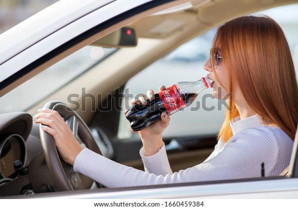 Kyiv, Ukraine - October\
14, 2019: Young woman driver drinking Coca-Cola soda drink while\
driving her car.
