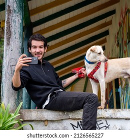 La Estrella, Antioquia, Colombia - February 13 2022: A Bearded Man Dressed In Black Takes A Picture With His Phone While Holding His Dog On A Leash