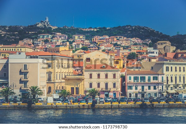 LA MADDALENA, ITALY. August 08, 2018:\
Port of La Maddalena in Italy. Colorful buildings on a hill, sea\
and numerous cars on the seafront. Blue\
sky.