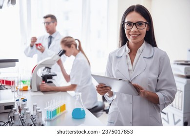 At the lab. Beautiful medical doctor is holding a digital tablet, looking at camera and smiling, her colleagues are working in the background - Powered by Shutterstock