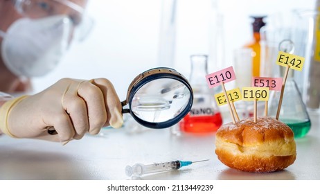 In the laboratory assistant's hand is a magnifying glass, on the laboratory table is a donut decorated with tablets with the names of E. Food Laboratory additives.