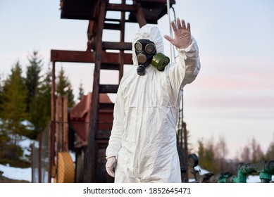 Laboratory Expert Wearing White Coverall, Gloves And A Gas Mask Working In Oil Field, Showing Stop Sign, Oil Pump Jack On Background, Concept Of Emission Of Oil Industry