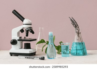 Laboratory glassware and microscope with ingredients for preparing natural cosmetics on white table near pink wall