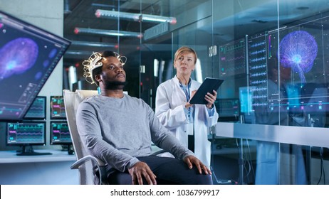 In Laboratory Man Wearing Brainwave Scanning Headset Sits in a Chair while Scientist with Tablet Computer Supervises Process. Monitors Show EEG Reading and Graphical Brain Model.