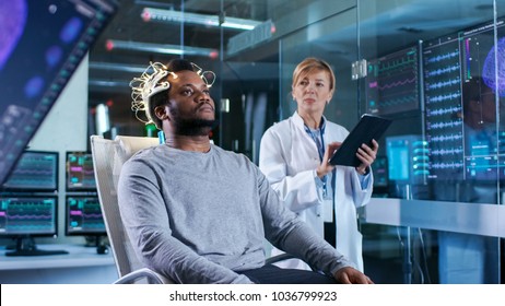 In Laboratory Man Wearing Brainwave Scanning Headset Sits in a Chair while Scientist with Tablet Computer Supervises Process. In the Modern Brain Study/ Neurological Research Laboratory.