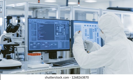 In Laboratory Over the Shoulder View of Scientist in Protective Clothes Holding Test Tube and Doing Research on Personal Computer. Modern Manufactory Producing Semiconductors and Pharmaceutical Items - Powered by Shutterstock