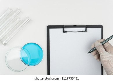 laboratory scene, the scientist holding a pen, watch glass, the test tubes and a clipboard on the white table. - Powered by Shutterstock