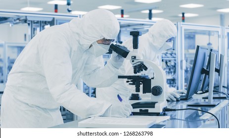 In Laboratory Scientist in Protective Clothes Doing Research, Using Microscope and Writing Down Data. Workers Working on a Modern Manufactory Producing Semiconductors and Pharmaceutical Items. - Powered by Shutterstock