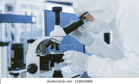 In Laboratory Scientist in Protective Clothes Doing Research Uses Microscope and Personal Computer. Modern Manufactory Producing Semiconductors and Pharmaceutical Items.