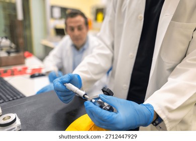 Laboratory technician using a precision micrometer to measure a metal component. Close-up of hands in blue gloves performing accurate dimensional inspection in a research or engineering lab.