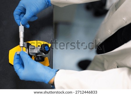 Laboratory technician using a precision micrometer to measure a metal component. Close-up of hands in blue gloves performing accurate dimensional inspection in a research or engineering lab.