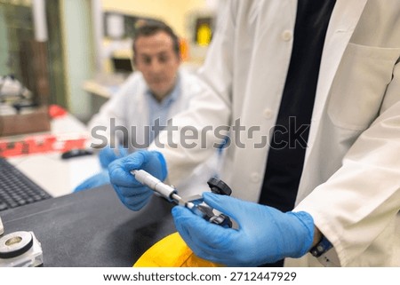 Laboratory technician using a precision micrometer to measure a metal component. Close-up of hands in blue gloves performing accurate dimensional inspection in a research or engineering lab.