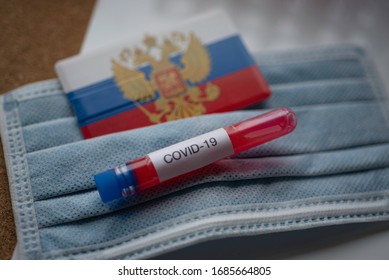 Laboratory Tube Test With Blood With COVID - 19 Label Next To Medical Mask And Blurred Flag Of Russia (Russian Federation). Coronavirus Blood Test And Vaccine Concept.  