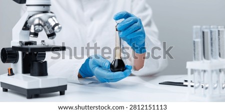 Laboratory workspace with microscope and gloved hands holding a volumetric flask of dark oil for scientific testing and analysis.