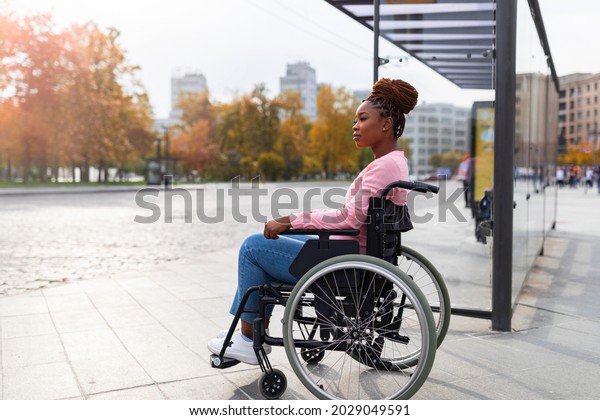 Lack of public transport for disabled people. Young\
black paraplegic woman in wheelchair feeling upset, waiting on bus\
stop in autumn, cannot board vehicle suitable for handicapped\
persons, copy space
