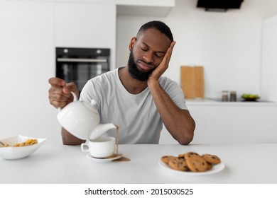 Lack Of Sleep Concept. Portrait of tired young African American man sleeping while sitting at dining table in kitchen, holding kettle pouring coffee away from cup spilling hot drink on desk