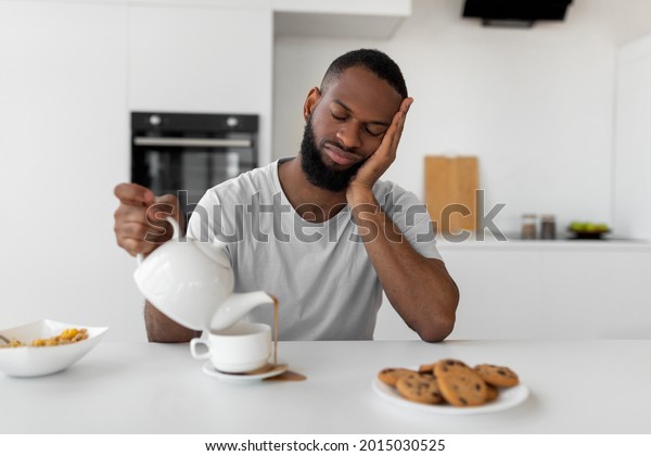 Lack Of Sleep Concept. Portrait of tired young\
African American man sleeping while sitting at dining table in\
kitchen, holding kettle pouring coffee away from cup spilling hot\
drink on desk