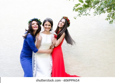 Ladies in blue and red gowns stand around bride posing before white wall
