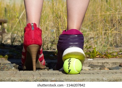 Ladies Legs, One Foot In The Court Shoe And The Other Foot In A Sports Shoe And A Tennis Ball With The Inscription  2018.