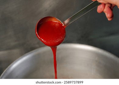 Cucharón de salsa de tomate vibrante: un primer plano de un cucharón de plata vertiendo salsa de tomate roja espesa y rica en una olla de acero inoxidable, ilustrando la preparación de un plato culinario.