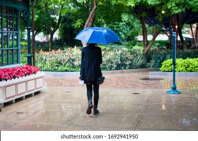 A Lady In All Black Clothes Walking Under A Light Drizzle While Holding A Small Blue Umbrella.