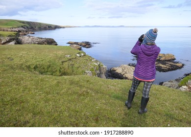 A lady in black boots ,winter clothes and bobble hat looks with binoculars at a coastline landscape of rocks and sea.Shetland.UK