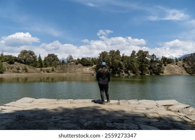 A lady in a blue cap and dark attire stands on a stone platform, gazing at a serene lake surrounded by rolling hills, lush green trees and snow-capped mountains. A vibrant blue sky at Deoria Tal lake. - Powered by Shutterstock